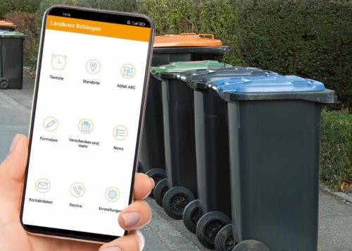 Abfall-App Handy mit der Abfall-App auf dem Bildschirm und Mülltonnen im Hintergrund.